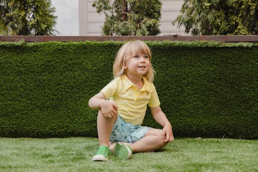 Happy child sitting on grass in a backyard setting with vibrant greenery.